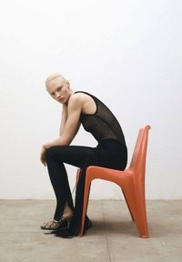Zwarte mesh top met franjes, zwarte slim-fit broek en patroon schoenen. Model zit op een oranje plastic stoel tegen een witte muur.