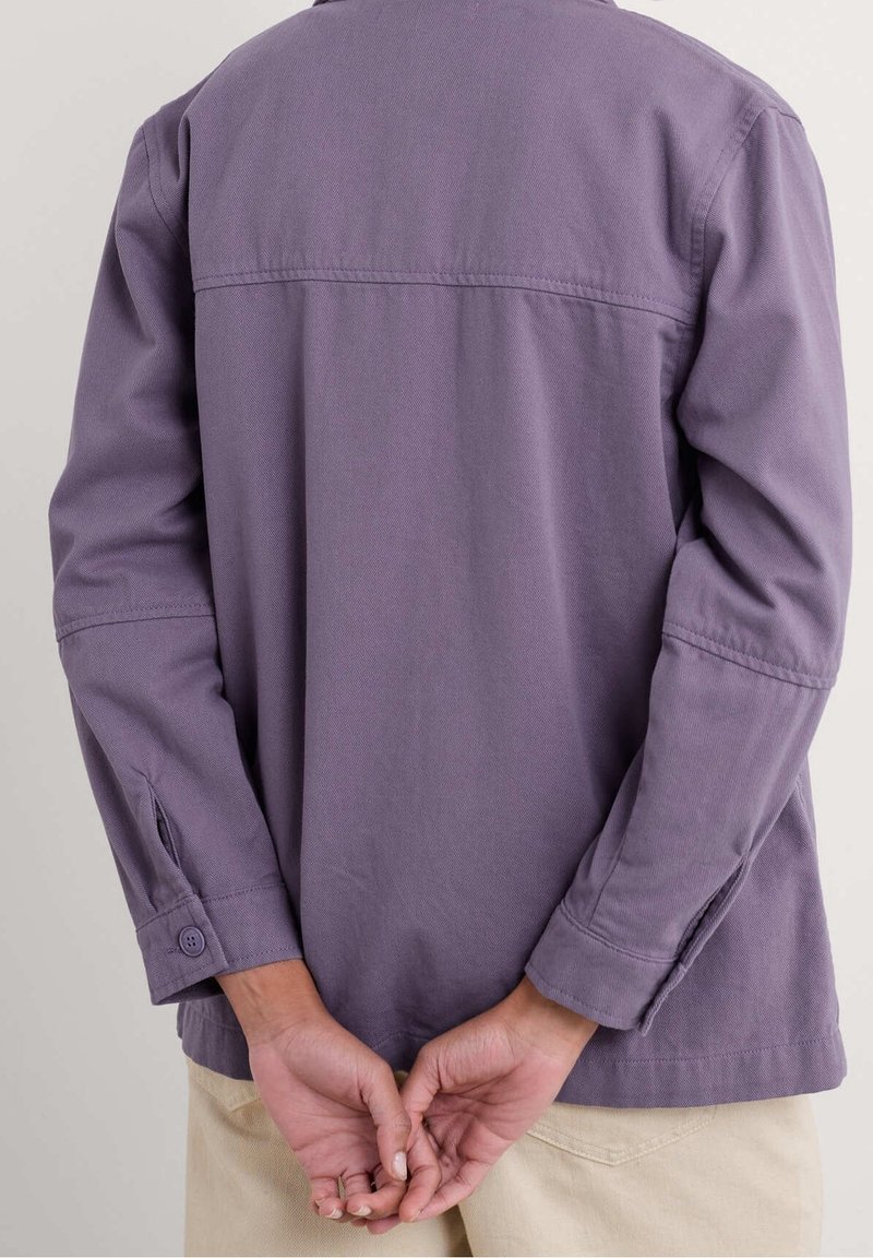 Fioletowa koszula z długim rękawem, zapinana na guziki przy mankietach, o prostym kroju i teksturowanej tkaninie, z panelem naramiennym z tyłu. Ręce złożone z tyłu.