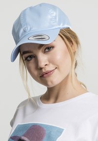 Lyseblå baseballcap lavet af glinsende materiale, med en bueformet skygge og et mærke på forsiden. Båret sammen med en simpel hvid T-shirt.