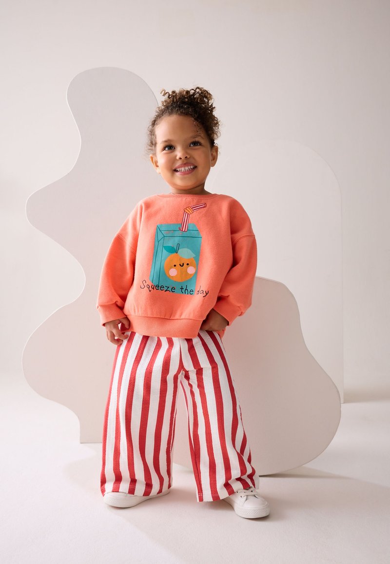 Naeratav laps, lokkis juustega, oranži pusa ja mahlapakiga, millel kiri "Pigista päev täis", punase-valge triibulise laiade pükstega ja valgete kingadega.