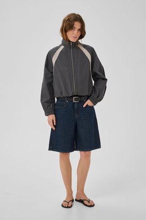 Žena u tamnoj jakni sa zatvaračem i bež detaljima, širokim traper šortsem, crnim remenom i crnim japankama stoji ispred jednobojne pozadine.