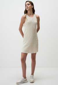 Gebreide beige jurk met een textuur, mouwloos ontwerp en een bescheiden lengte, gecombineerd met wit en beige sneakers.