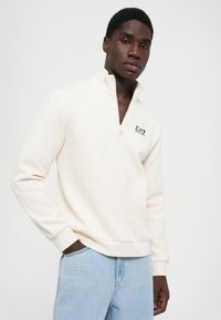 Ung mann iført en hvit quarter-zip hettegenser med EA7-logo og lyseblå jeans, stående med den ene hånden i lommen mot hvit bakgrunn.