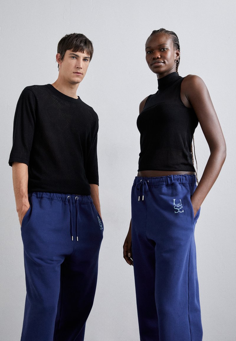 Schwarzes kurzärmliges Mesh-Oberteil und schwarzes ärmelloses Oberteil mit hohem Kragen, kombiniert mit blauen Sweatpants mit Zugband und gesticktem Logo.