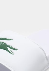 Sandale blanche à semelle plate avec un logo de crocodile vert, dotée d'une texture lisse et d'une large sangle. Design simple sans motifs ou accents supplémentaires.