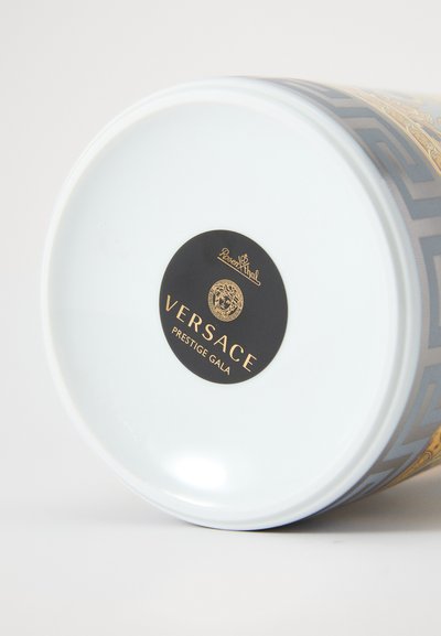 Recipiente cerâmico com base branca brilhante. Apresenta um padrão geométrico dourado e azul. Rótulo preto com a marca "Versace Prestige Gala".