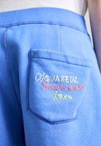 Pantalones cortos de sudadera azul claro con un bolsillo parche y texto bordado multicolor: "DSQUARED2 SUMMER LOVERS XOXO". Textura de tela suave.