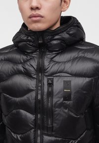 Modèle masculin portant une doudoune noire brillante avec un col haut et une poche poitrine zippée, affichant le logo Blauer sur la poitrine.