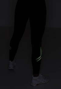 Czarne, refleksyjne legginsy sportowe o dopasowanym kroju, z świecącymi akcentami po bokach, zestawione z białymi, teksturowanymi sneakersami.