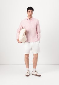 Lyserødstribet skjorte med knapper, hvide teksturerede shorts, en cremefarvet rygsæk og beige sneakers med brune accenter. Modellen står foran en hvid baggrund.