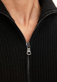 Svart stickad zip-up tröja med hög krage och silverfärgad dragkedja. Texturen är ribbad och tyget verkar tjockt och mysigt.