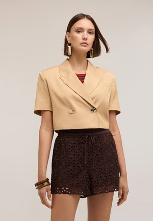 Vrouw met een beige cropped double-breasted blazer, zwarte kanten shorts, gouden opvallende oorbellen en bruine armbanden, staand tegen een neutrale achtergrond.