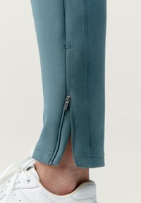 Modré športové nohavice s detailom na zipsu pri členku, vyrobené z mäkkej tkaniny, skombinované s bielymi teniskami, ktoré odhaľujú časť členka.