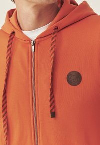 Ritsvest in oranje stof met een gestructureerde afwerking. Voorzien van zwarte gestreepte trekkoorden en een ronde bruine patch met berendesign op de borst.
