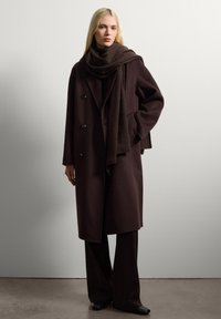 Brown wool coat with a double-breasted design, black buttons, and large lapels. Accompanied by a matching dark scarf and wide-leg pants.