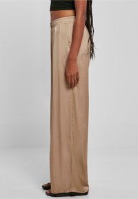 Beige, wijde satin broek met een elastische tailleband, een gladde textuur en zijkant detailing, gezien vanaf de zijkant.