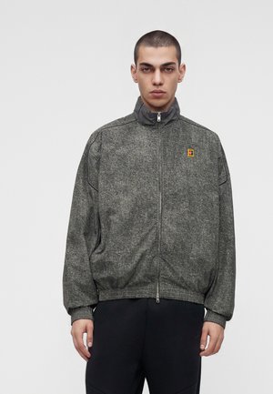 Jeune homme aux cheveux courts portant une veste grise zippée avec un petit patch coloré sur la poitrine, debout devant un fond clair uni.