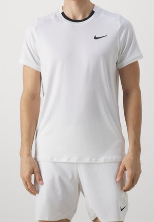 Mann trägt weißes Nike-Sportshirt und Shorts, steht mit entspannten Armen seitlich am Körper vor einfachem Hintergrund.
