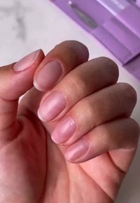 Delikatnie przystrzyżona dłoń z naturalnie wyglądającymi, jasnoróżowymi paznokciami o gładkim wykończeniu i subtelnym połysku, na miękkim tle.