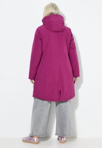 Woman with blonde hair wearing a long magenta hooded coat, wide gray pants, and purple metallic sneakers facing a white wall.