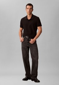 Man staat naar voren gericht, gekleed in een zwart poloshirt, zwarte riem, donkergrijze jeans en zwarte schoenen, tegen een eenvoudige lichtgrijze achtergrond.