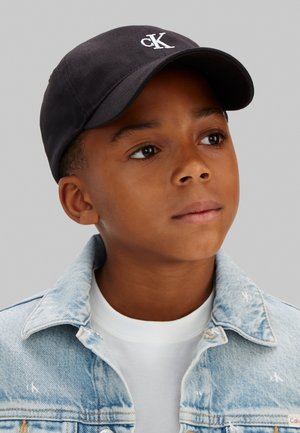 Junge mit schwarzer Calvin Klein Mütze, weißem T-Shirt und hellblauer Jeansjacke, der leicht nach oben und zur Seite gegen einen einfarbigen Hintergrund blickt.