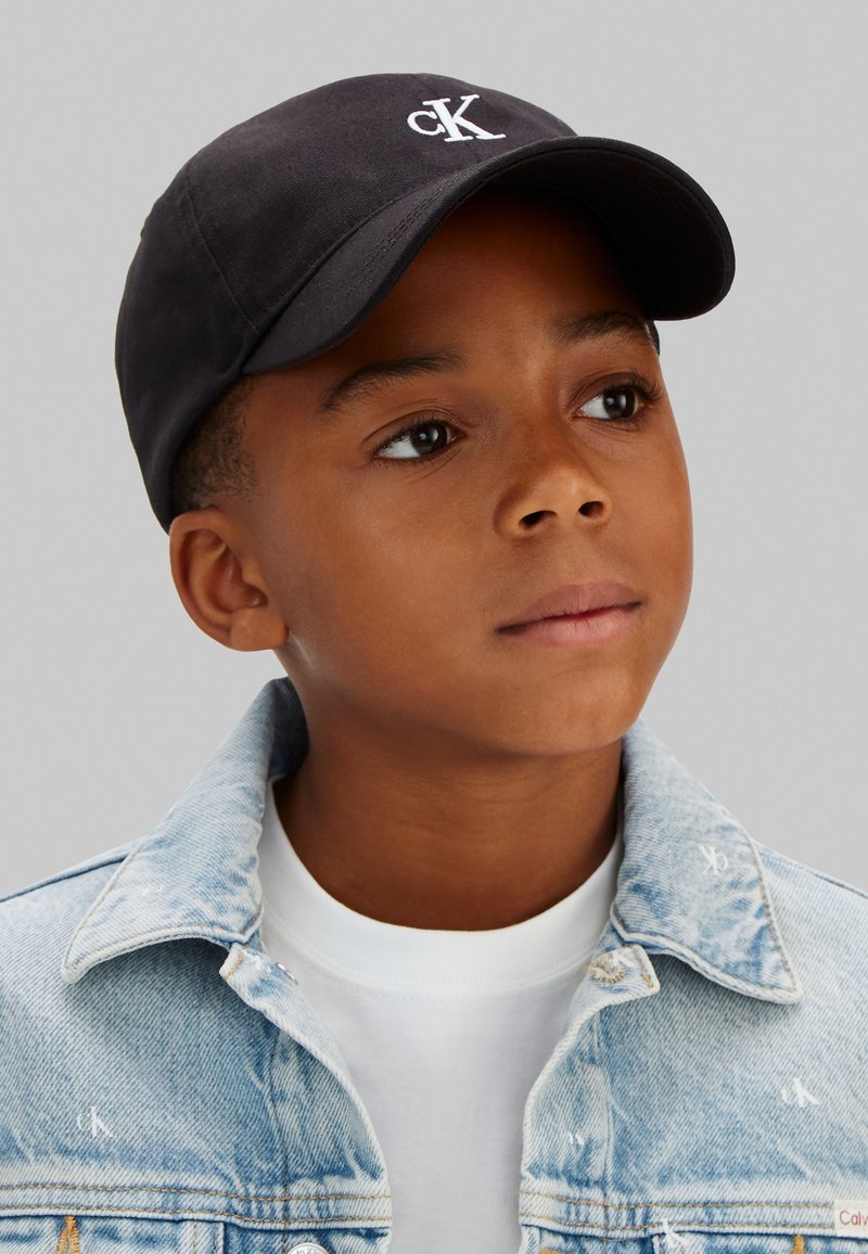 Fekete Calvin Klein sapkát, fehér pólót és világoskék farmerdzsekit viselő fiú, aki enyhén felfelé és oldalra néz egyszínű háttér előtt.