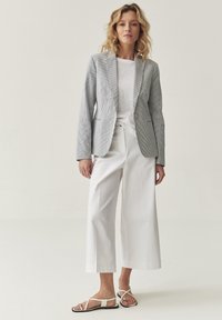 Blazer grigio a righe su una camicia bianca annodata, abbinato a pantaloni bianchi larghi e sandali flat. L'outfit combina diverse trame e colori.