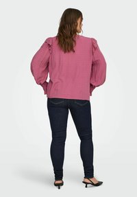 Kobieta stoi tyłem, ubrana w teksturowaną różową bluzkę z falbanami na ramionach, ciemne dopasowane dżinsy i czarne buty na niskim obcasie.