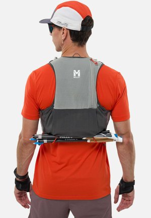 Homme en chemise orange et casquette portant un sac à dos gris avec des bâtons de trekking attachés, vu de dos.