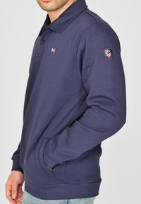 Tummansininen pusero, jossa on kaulus, vasemmassa hihassa on paikka ja sivutaskut. Valmistettu pehmeästä, teksturoidusta kankaasta.