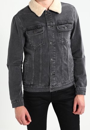 Uomo con indosso una giacca di jeans grigio scuro con colletto in shearling crema e pantaloni neri, su sfondo semplice.