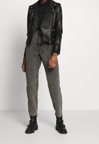 Personne portant une veste de biker en cuir noire, un col roulé noir, un jean taille haute gris délavé et des bottines noires à boucle sur un fond uni.