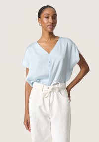 Blusa azzurro chiaro a maniche corte con scollo a V, caratterizzata da una texture plissettata, abbinata a pantaloni bianchi a vita alta con texture.