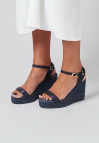 Schwarze Keilsandalen mit einer strukturierten, geflochtenen Plattform, ausgestattet mit zwei Knöchelriemen und einem metallischen Glanz. Offenes Toe-Design mit schwarzem Nagellack.