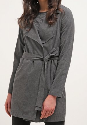 Kobieta ubrana w szary płaszcz o długości do połowy uda, z długimi rękawami i paskiem związanym w talii, na ciemnoszarym topie.