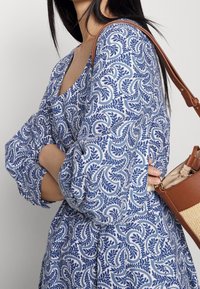 Blau-weißer, gemusterter Kleid mit Puffärmeln und V-Ausschnitt. Eine braune Schultertasche mit geflochtenem Akzent wird seitlich gehalten.