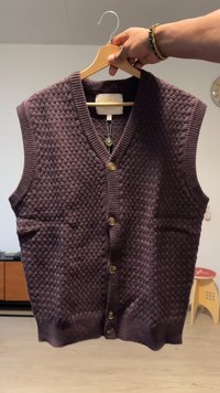 V-neck, sleeveless cardigan in dark purple with a textured pattern, featuring five buttons and two front pockets. Wood hanger visible.