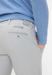 Man met lichtgrijze broek en lichtblauwe trui, met de rechterhand in de zak, gezien van de onderrug tot halverwege het bovenbeen.