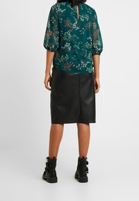 Blouse florale teal avec des manches transparentes et un dos en trou de serrure, associée à une jupe crayon en cuir noir et des bottines noires à boucles.
