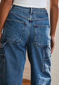 Pantaloni cargo din denim albastru cu talie înaltă, având buzunare mari la spate și buzunare laterale. Țesătura are un finisaj texturat și decolorat.