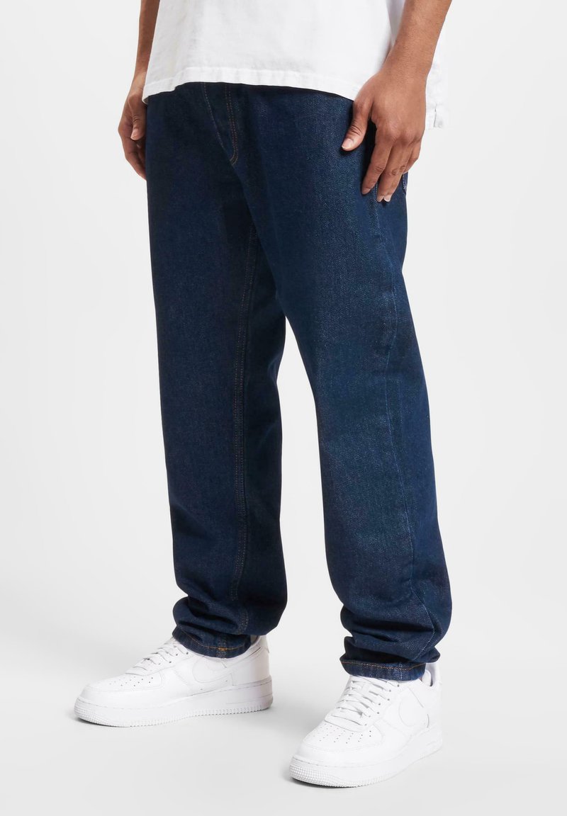 Dark blue denim jeans with a relaxed fit, featuring visible stitching, side pockets, and worn-out detailing on the front. Paired with white sneakers.