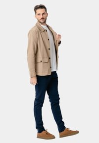 Beige jakke med krave og knapper, to forlommer, båret over en hvid skjorte, parret med marineblå bukser og brune sko.