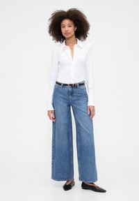 Bela srajca z dolgimi rokavi na gumbe, modre široke kavbojke s črnim pasom in črna balerinka. Tkanina deluje gladko in sproščeno.