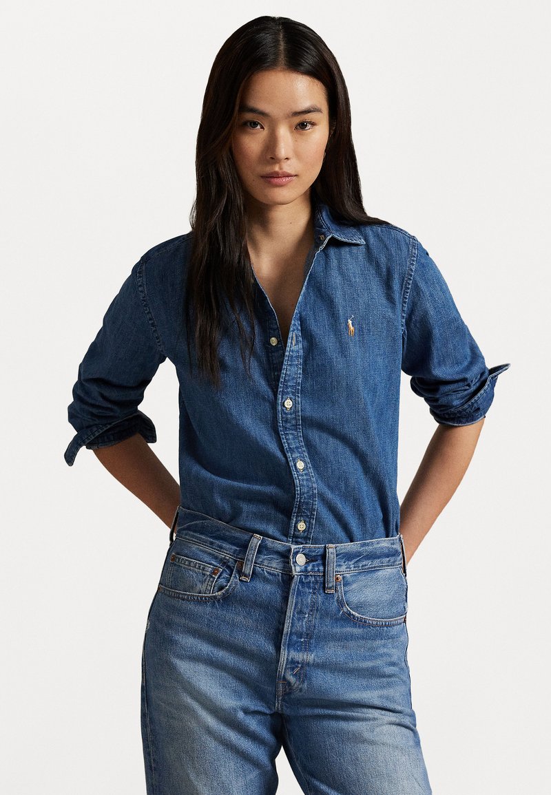 Jovem mulher com cabelo escuro e comprido, vestindo uma camisa de denim azul e jeans azuis, com as mãos apoiadas nas costas.