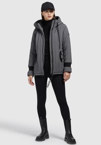 Gray quilted jacket with a hood, black accents, zipper pockets, paired with black leggings and chunky black boots. Casual, functional design.