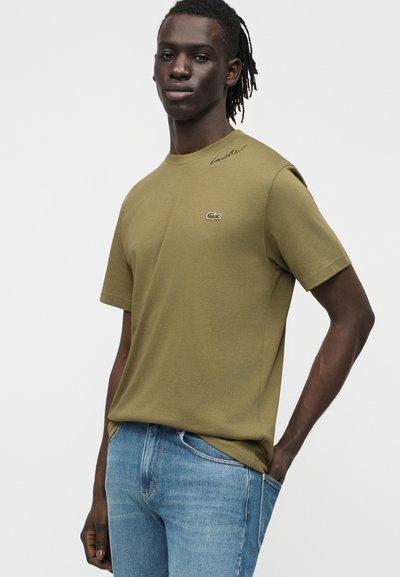 Млад мъж с тъмна кожа и дредове носи оливенозеленa тениска на Lacoste и сини дънки, стои с една ръка в джоба срещу бял фон.