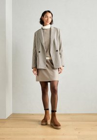 Blazer de cuadro de doble botonadura, suéter beige, falda beige ajustada, medias negras transparentes y botas marrones con suela de plataforma.