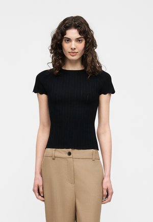 Γυναίκα με καστανά σγουρά μαλλιά που φορά στενό μαύρο κοντομάνικο τοπ και μπεζ ψηλόμεσα παντελόνια, στέκεται μπροστά σε ένα απλό λευκό φόντο.