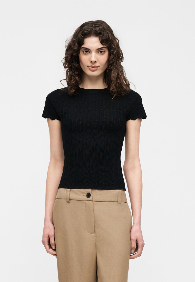 Γυναίκα με καστανά σγουρά μαλλιά που φορά στενό μαύρο κοντομάνικο τοπ και μπεζ ψηλόμεσα παντελόνια, στέκεται μπροστά σε ένα απλό λευκό φόντο.